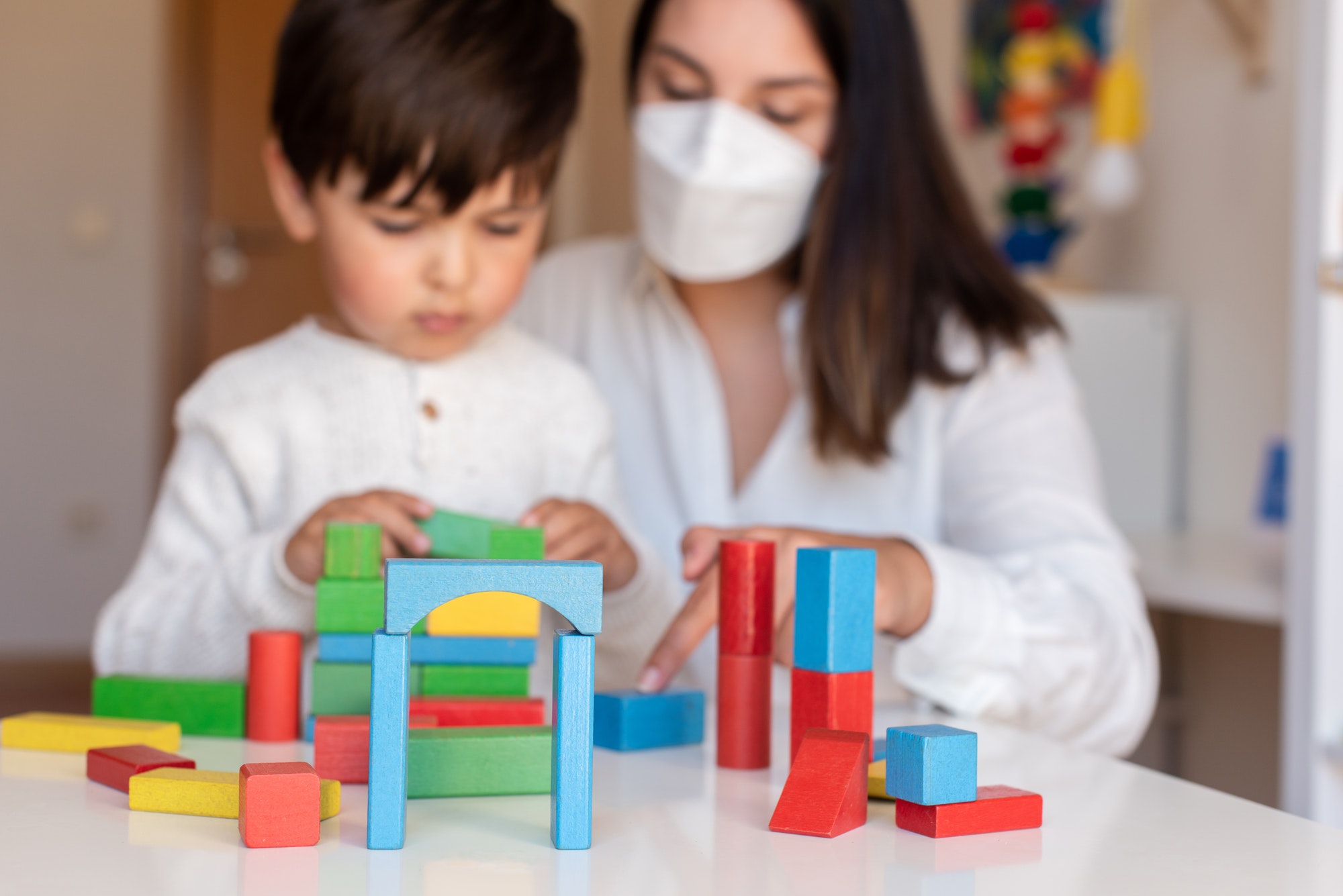 Kid playing with wood blocks and teacher educador help using face mask for coronavirus pandemic.